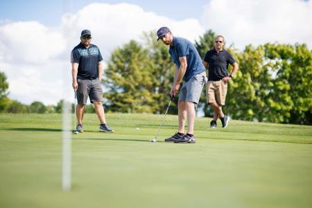Three men playing golf on a putting green, with one man lining up and making a putt while the other two watch nearby on the green.