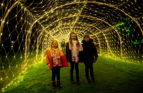 Three young children outdoors facing the camera beneath a tunnel of fairy lights