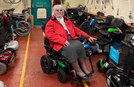 A female in a mobility scooter smiling towards the camera in front of lines of mobility scooters and wheelchairs