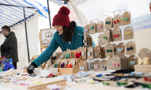 A woman wearing a red wooly hat and a blue coat manning an arts and crafts stall