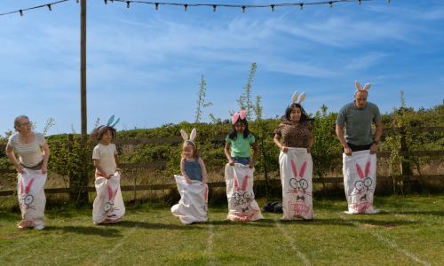 A line of children and adults wearing bunny ears and standing in white easter bunny themed sacks at the start of a race track.