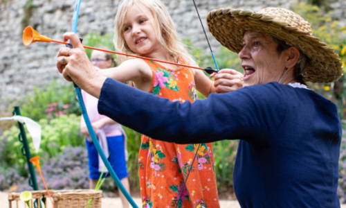 A woman wearing a straw hat teaching a girl how to do archery