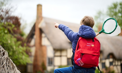 A boy with a red backpack on pointing towards Anne Hathaway's Cottage and holding a green net in the other hand