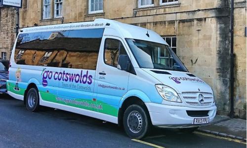 The Go Cotswolds branded minibus parked outside a honey coloured building on a Cotswolds street