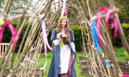 A young female dressed in Tudor costumer walking through an arch of sticks decorated with colourful ribbons