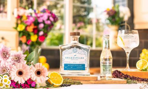 A bottle of Mother's Ruin Gin by Shakespeare Distillery set on a table next to a bottle of tonic and a glass of gin and tonic surrounded by summer flowers