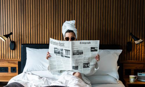 A woman sitting in bed wearing sunglasses, a white dressing gown with a white towel turban on her head reading the Telegraph newspaper.