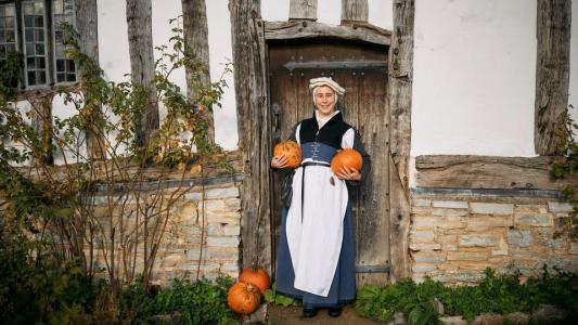 A lady in Tudor costume standing in front of a cottage holding a pumpkin in each hand