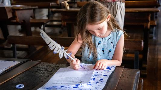 A young girl seated at a desk in an Elizabethan schoolroom writing with a quill