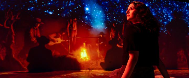 A woman with curly hair sits on a cushioned bench in a dark, immersive gallery space, lit by red and blue light. Behind her, a large panoramic screen shows a night sky filled with bright stars above silhouettes of people gathered around a campfire, some playing guitar.