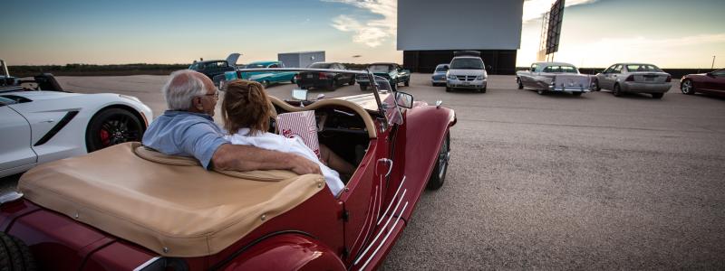 A couple sits in a vintage car in front of a movie screen at Stars & Stripes Drive-In Theater
