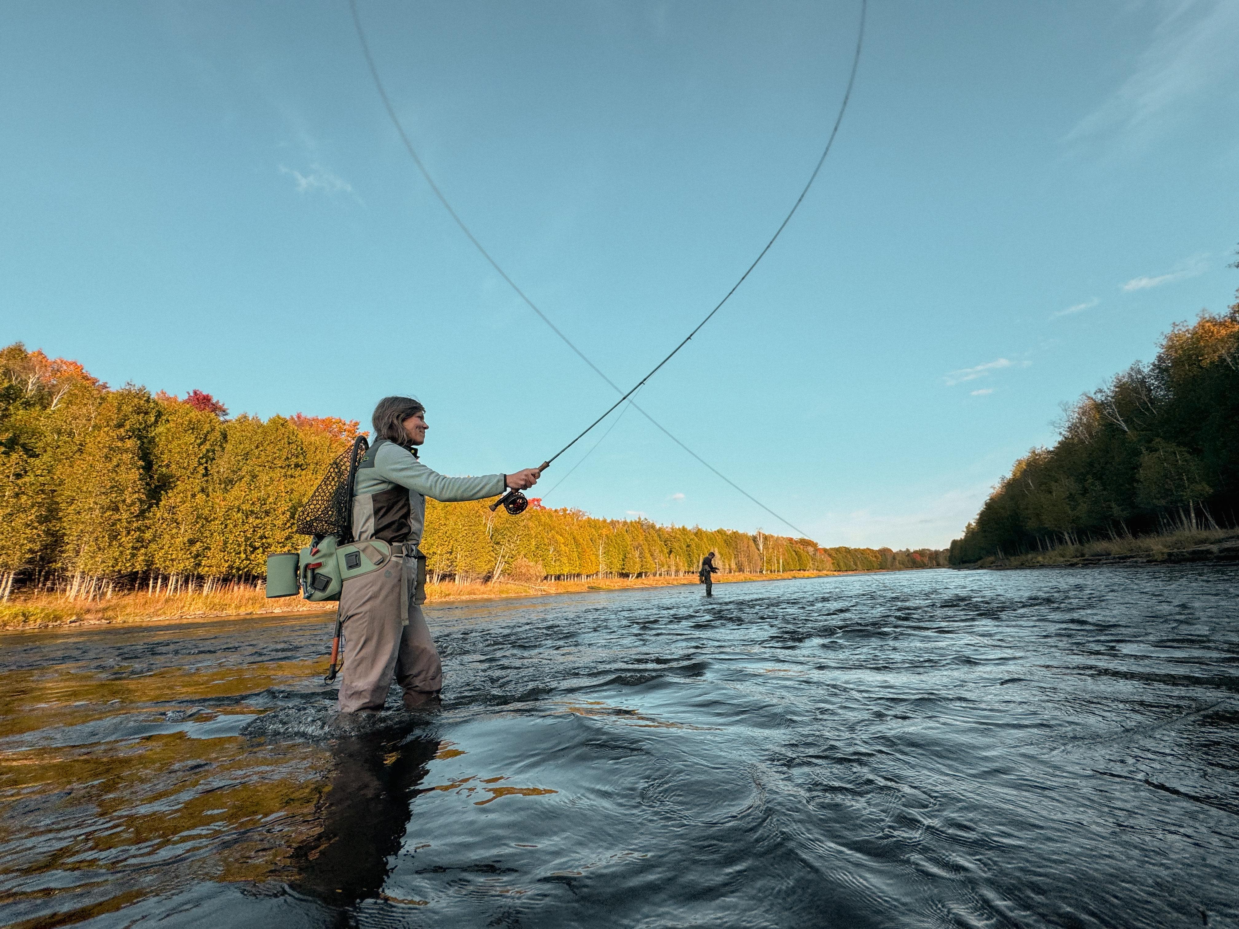 A woman fly-fishes in a river near Escanaba surrounded by autumn trees under a clear blue sky. The water reflects the warm hues, creating a serene, focused atmosphere.