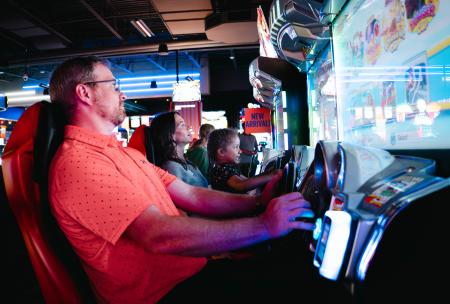 A father sits in his own chair beside a mother and daughter who are sharing a chair while the family plays an arcade game at Dave & Buster’s.