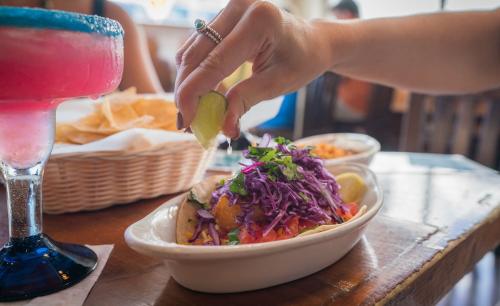 Woman squeezing lime on top of a taco piled high with toppings of cabbage and cilantro - next to her is a pink and blue frozen margarita and a basket of chips in the background