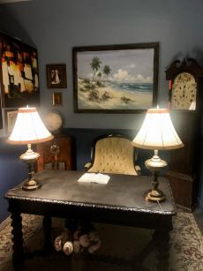 A dimly-lit room is filled with a wooden desk with two lamps on it, a regal desk chair, and art on the walls.