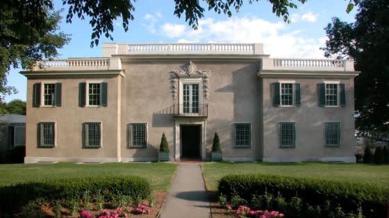 A historic two-story beige building with green shutters and ornate central entrance flanked by two symmetrical wings, set in landscaped gardens under a clear sky.