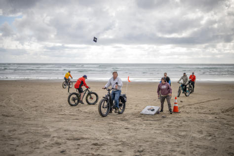 People riding their fat tire bikes on the beach tossing corn hole bags towards a board.