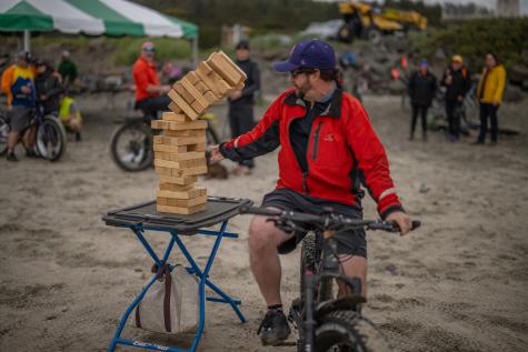 A person attempting to remove a Jenga block while riding their fat tire bike on the beach, topples the Jenga tower.