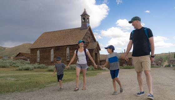 Family holding hands walking dirt roads of Bodie State Historic Park in front of old wooden church building