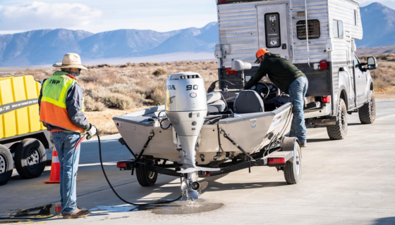 Inspectors checking and spray washing a trailered boat attached to a truck with mountains in the background