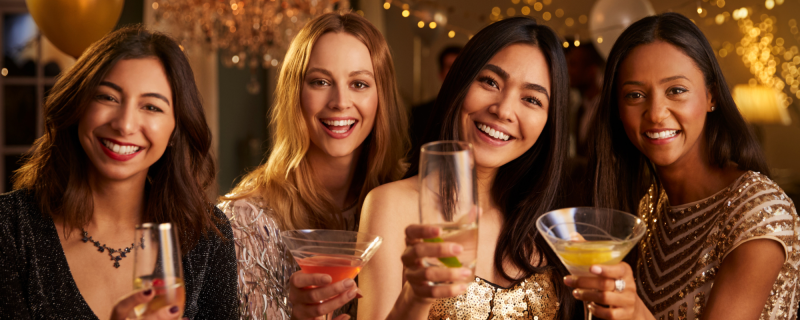 Four women hold up cocktails and smile with Christmas lights and decor in the background