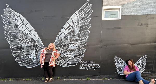 two women posing in front of a mural with white wings