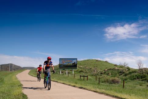Two people biking on 6th Avenue paved trail past Welcome to Golden sign and Fossil Trace Golf Course