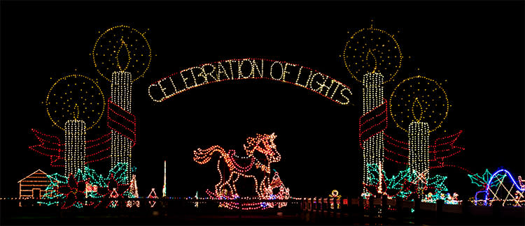 A lit up sign reading "Celebration of Lights" surrounded by tall lit candle lights with a rocking horse lit underneath is the entrance for Meadow Lights Christmas Light Display.