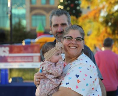 A woman holds a baby, smiling, with a man standing behind them. A festive outdoor setting features colorful decorations and people in the background.