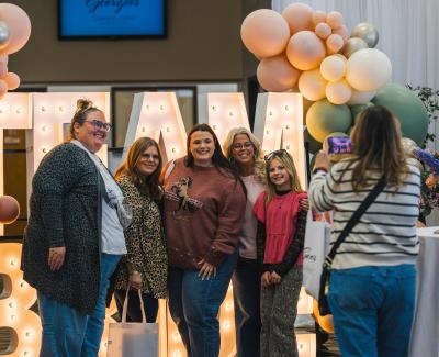 A group of five women and a girl pose together in front of a large illuminated "TEAM" sign, decorated with pastel balloons.