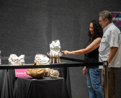 Two visitors examine a display of unique sculptures at an art exhibition. A sign reads "Christa G." in the background.