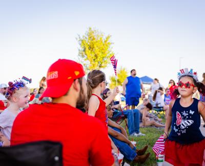 A crowd at a 4th of July event with a girl in a USA headband and star sunglasses.