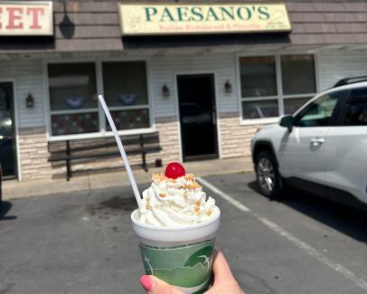 a hand holds a small ice cream sundae with whipped cream & a cherry on top in front of Paesano's Italian Restaurant & Pizzeria