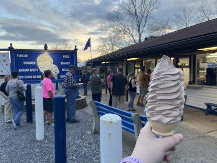 A hand holds a cone of chocolate soft serve ice cream in front of The Little Ice Creamer near Orbisonia, PA