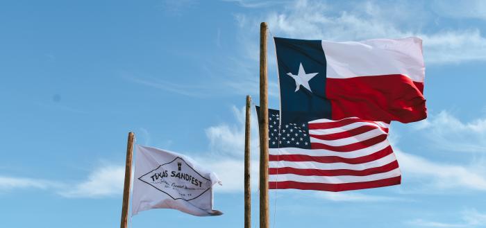 Three flags fly against a blue sky. One is the US flag, one is the Texas flag, and one is a white Texas SandFest flag