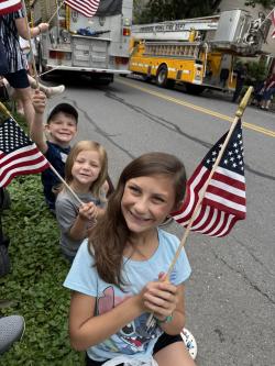 Picture of three children holding American flags while sitting on the curb of a sidewalk watching a July 4th parade.