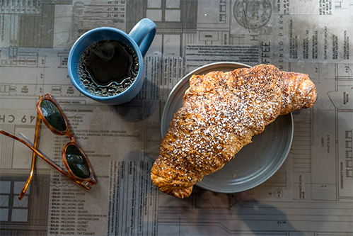 A croissant on a plate next to a cup of dark coffee and sunglasses.