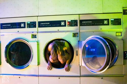 Performer crawling out of a washing machine at a laundry mat.