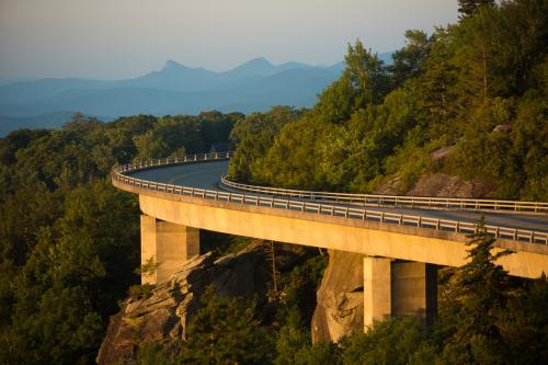 A bridge is held up by huge columns and hugs the curve of the mountain it navigates around. Blue mountain peaks can be seen in the background.