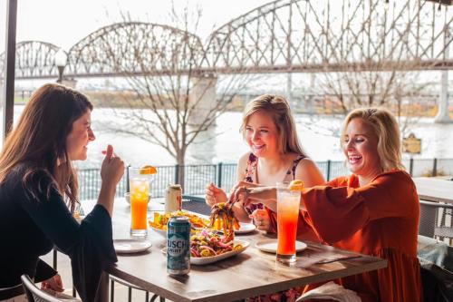 Three women eating and laughing at a patio table with an Ohio River bridge in the background.