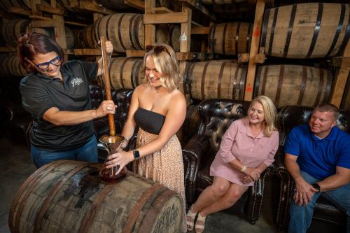 A young woman thieves bourbon from the barrel at Jeptha Creed Distillery as two adults watch.