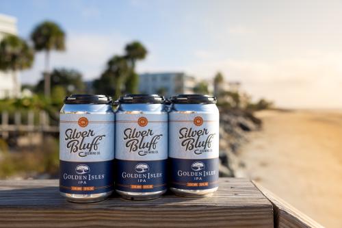 A six pack of Golden Isles IPA sits on a wooden railing in front a beach spotted with palm trees