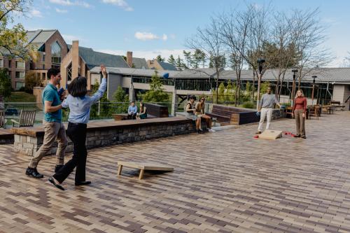 Four people are outdoors on a stone patio playing a game of cornhole.