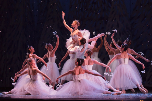 More than a dozen young girls dressed in white surround the Sugar Plum Fairy, also dressed in white in the center of the circle.