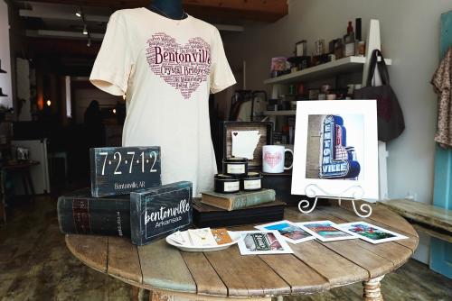 A product display at Blue Moon Bentonville featuring a Bentonville themed t-shirt, home decor, postcards, mug, and more.