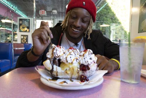 A man with a red had smiles and looks at a large banana split with whipped cream and cherries. His spoon is raised and just starting to dip into the ice cream. Blue booths and a strip on neon lights are seen in the background.