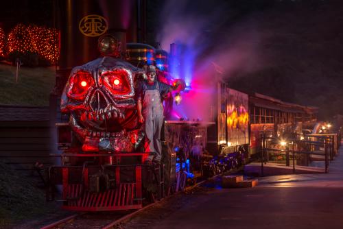A ghostly man stands on the front car of a train with the face of a skeleton, aglow with spooky lights.
