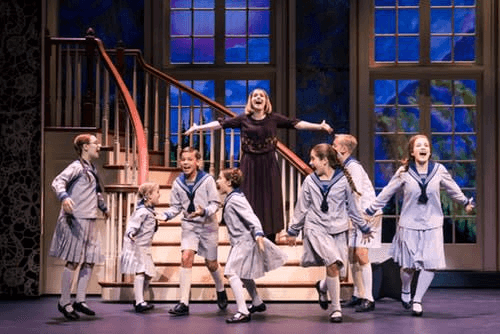 On a theatrical stage, a woman in a brown dress stands atop stairs, arms outstretched, surrounded by seven children in matching striped outfits, all in mid-action as if singing or dancing.