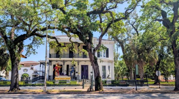 The Mobile Carnival Museum is a two story victorian style historic home painted white that sits behind tall oak trees