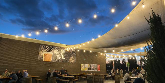 Photo shows beer garden with string lights, a sign that reads "Hella Local" and people sitting at tables drinking craft beer